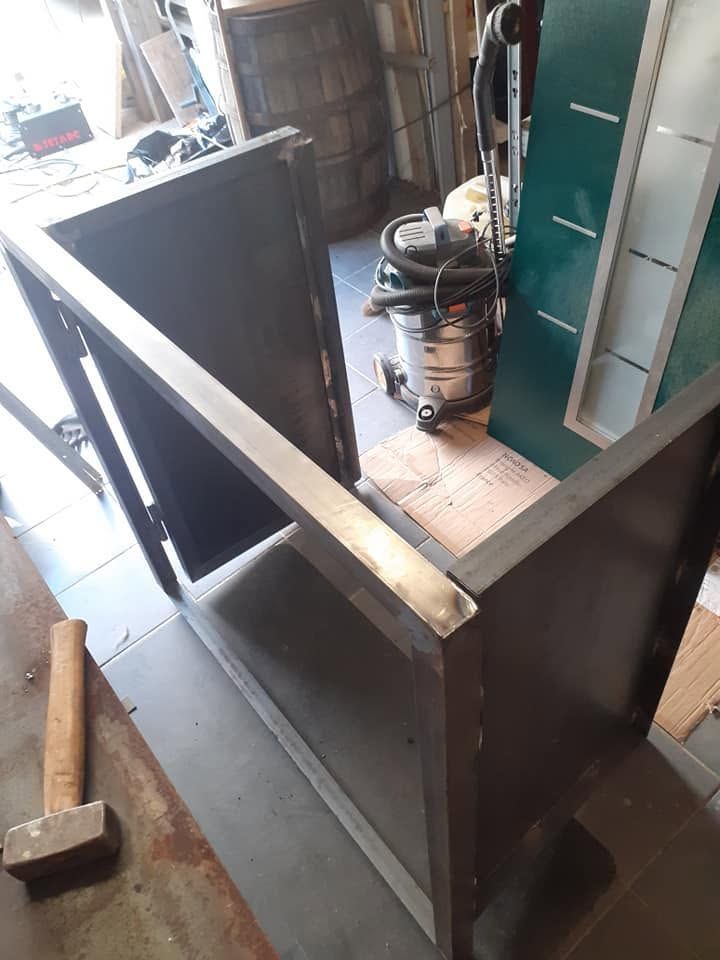 Boîtier en métal noir dans un atelier, avec un cadre métallique, une porte verte et un aspirateur posé dessus.