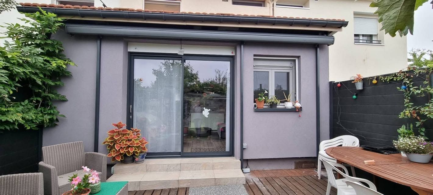 Patio arrière avec des portes-fenêtres coulissantes grises, des plantes en pot et un coin salon extérieur.