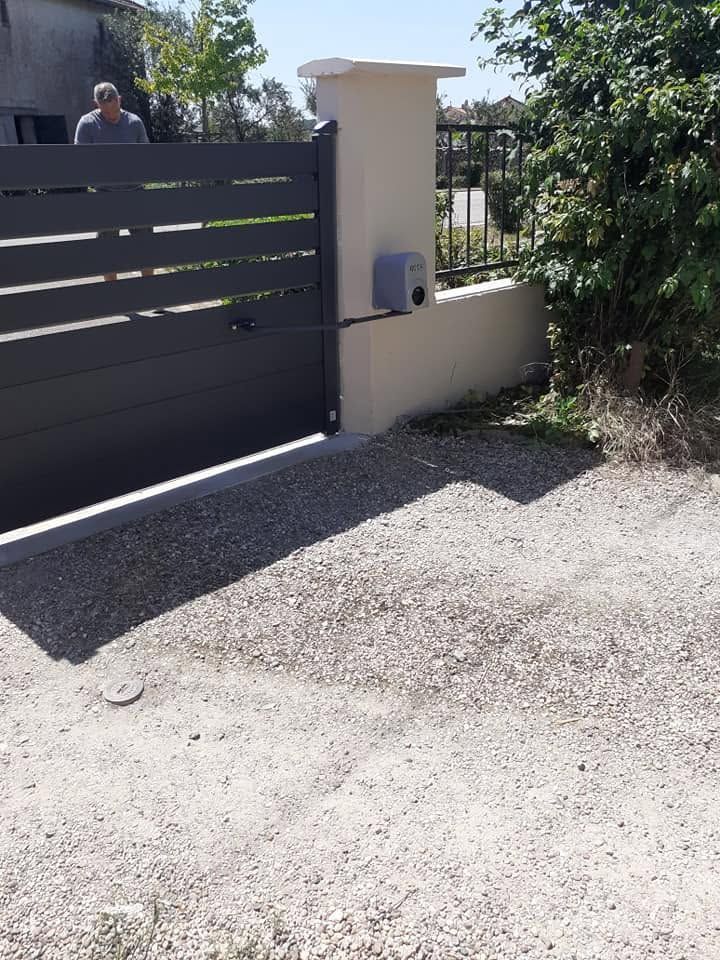 Entrée de garage avec portail sombre, pilier crème et allée en gravier bordée de verdure par une journée ensoleillée