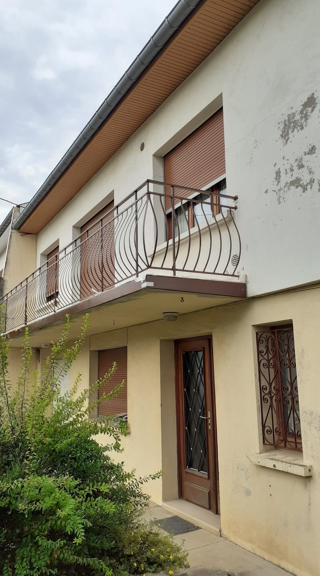 Maison à deux étages avec une façade beige, un balcon, des fenêtres à volets et une petite allée de jardin.