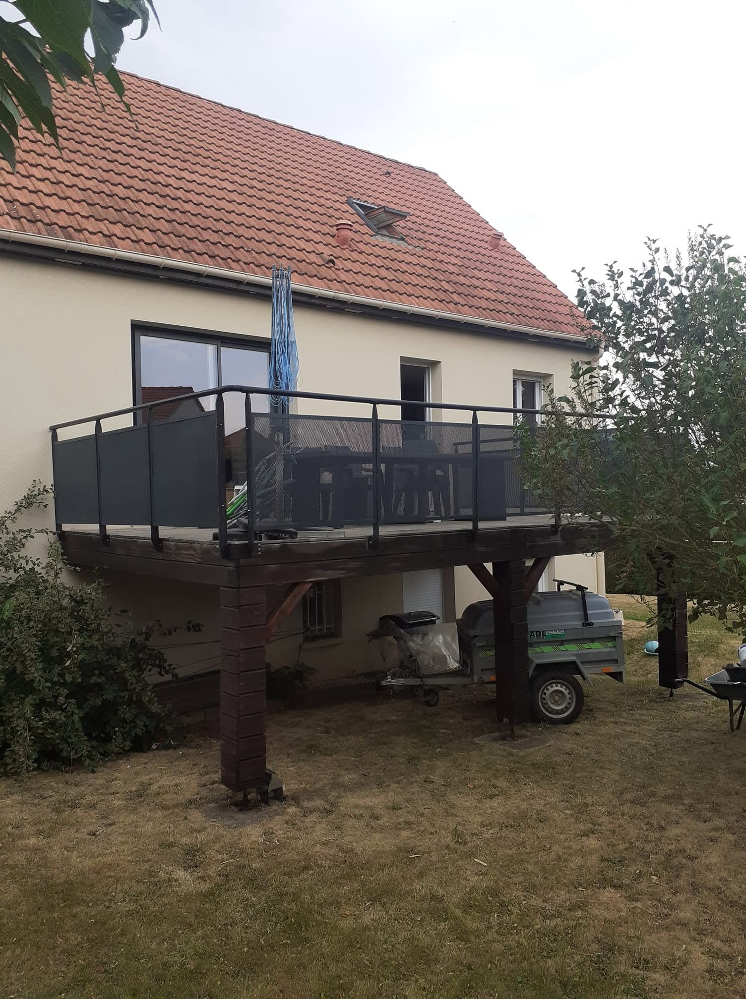 Maison avec une terrasse surélevée sur pilotis, dans une cour, avec un véhicule utilitaire garé dessous.