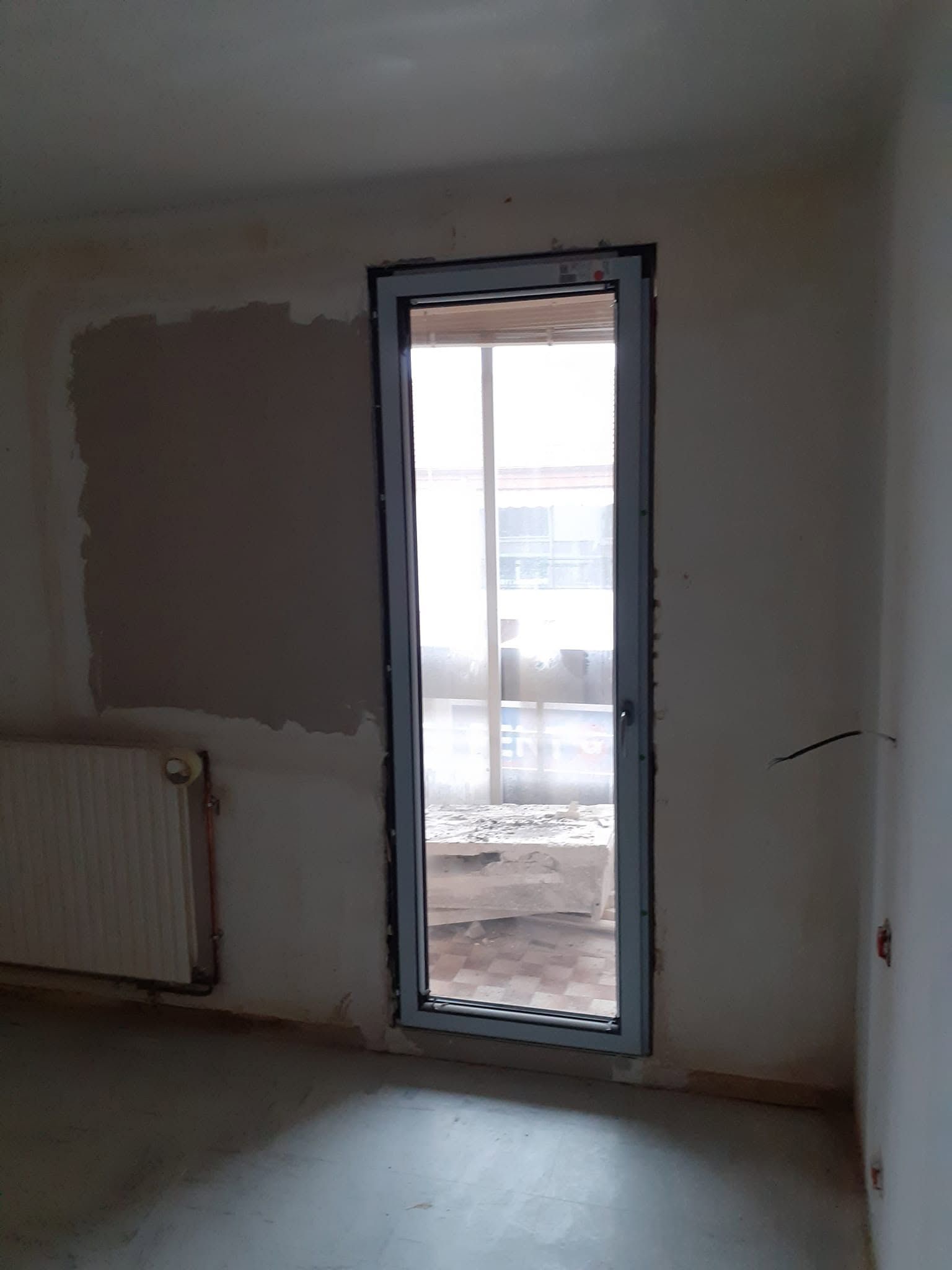 Pièce vide aux murs non finis, avec un radiateur et une porte vitrée donnant sur un balcon.