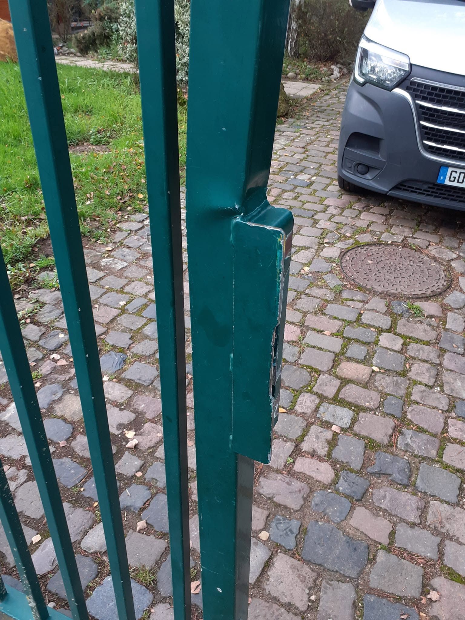 Loquet de portail métallique vert sur une allée pavée, avec une voiture grise garée à l’arrière-plan