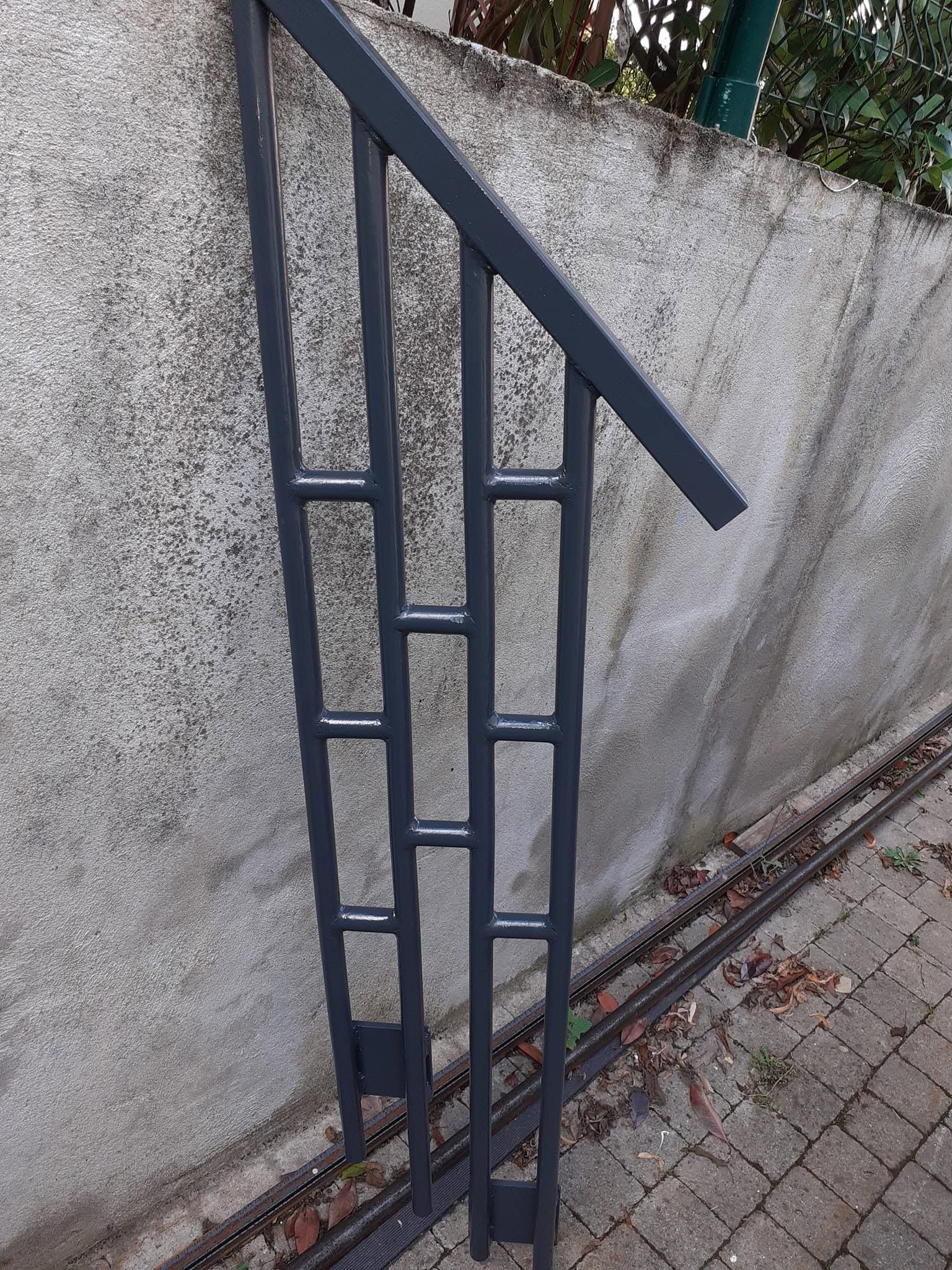Rampe en métal à barreaux verticaux longeant un escalier ou une rampe en béton, le long d’un mur et à côté d’un chemin pavé.