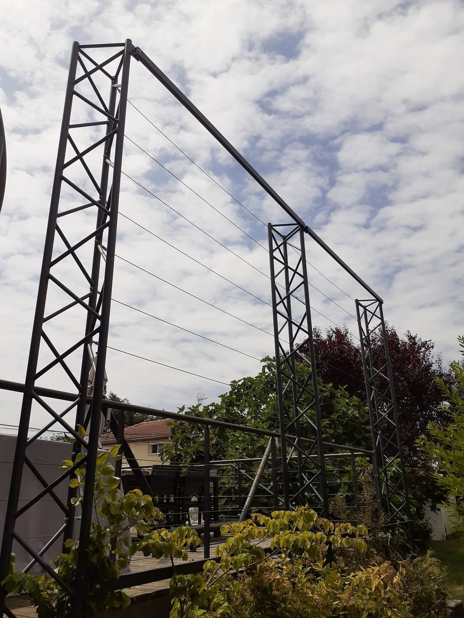 Passerelle suspendue avec des tours métalliques au-dessus d’un jardin, sous un ciel nuageux