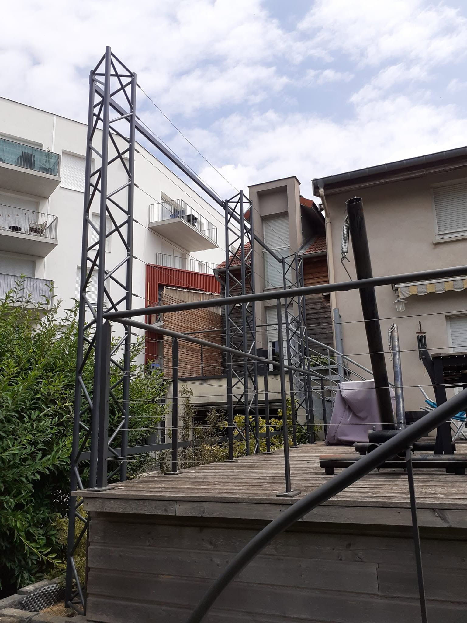 Petite terrasse dans un jardin avec un escalier en métal noir, une rambarde en bois.
