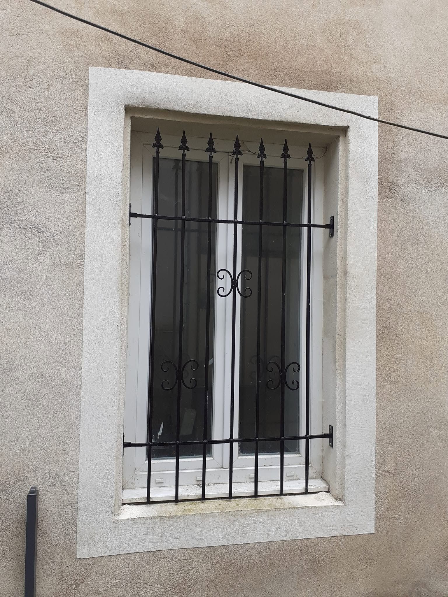 Fenêtre étroite à barreaux, encastrée dans un mur clair en stuc et dotée d’un cadre blanc.
