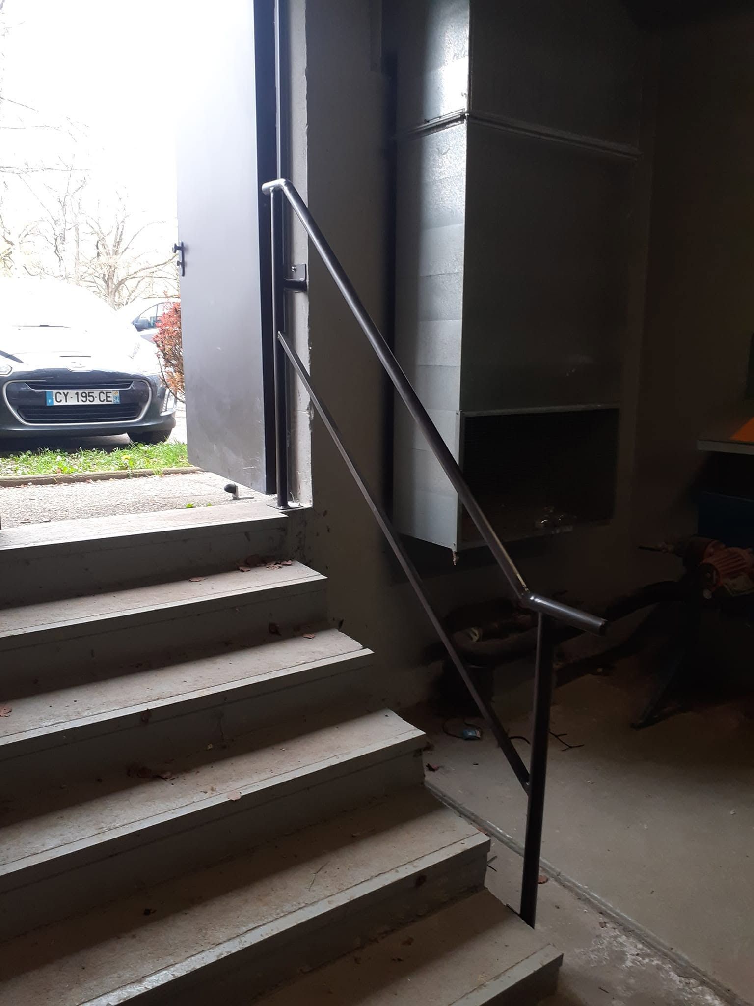 Cage d’escalier intérieure avec une rampe en métal noir, éclairée par la lumière du jour qui entre par une porte ouverte.
