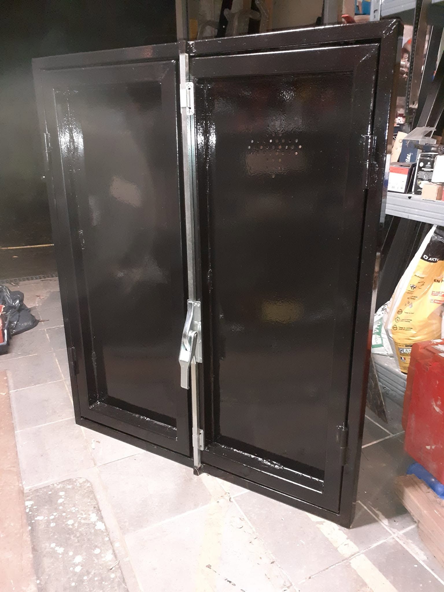 Armoire métallique noire à deux portes dans un atelier ou un magasin, partiellement ouverte sur un sol carrelé