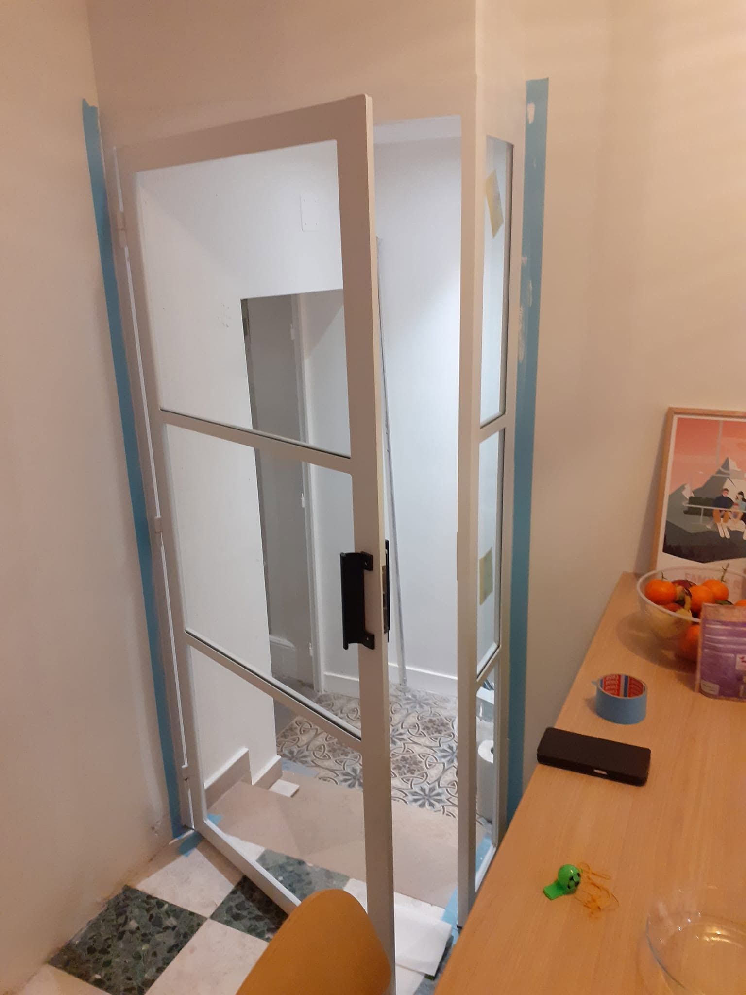 Porte vitrée ouverte avec un cadre bleu, dans une petite pièce à côté d’une table couverte d’objets éparpillés.