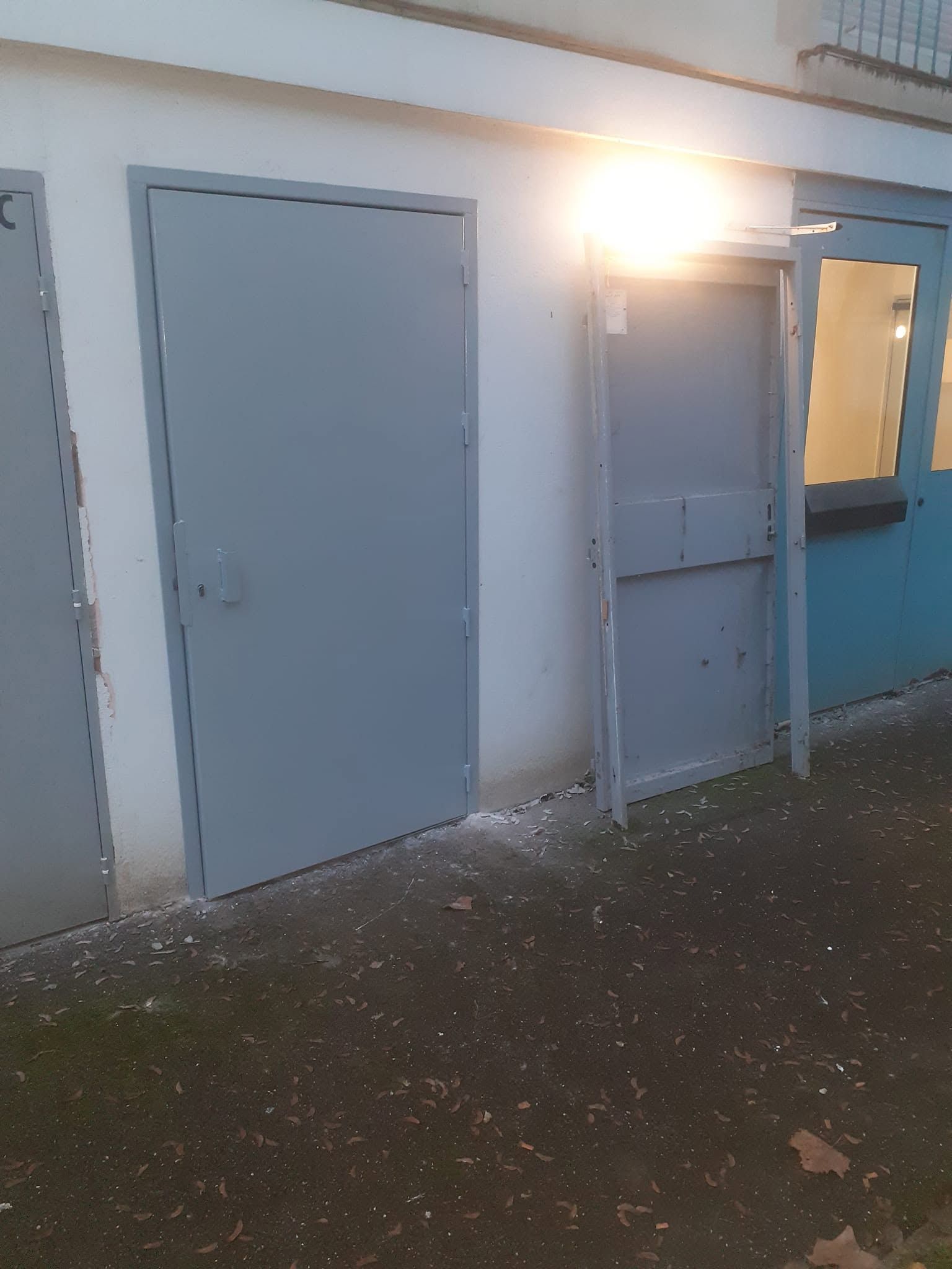 Extérieur d’un bâtiment avec des portes grises et une porte latérale bleue, baigné par une vive lumière du soleil.
