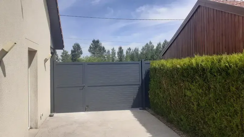 Allée bordée d’une maison beige et d’une haute haie verte, avec un portail métallique gris sous un ciel bleu