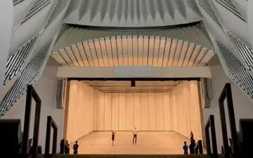 Vista interior de un escenario teatral con bailarines. Cortinas de colores claros forman el telón de fondo; el espacio está rodeado de elementos arquitectónicos.