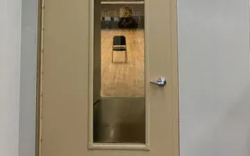 Puerta beige con ventana que da a una habitación con una silla. La habitación tiene suelo gris y paredes beige.