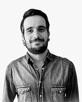 Hombre de cabello oscuro y barba, vestido con una camisa vaquera, sonriendo, sobre un fondo blanco.