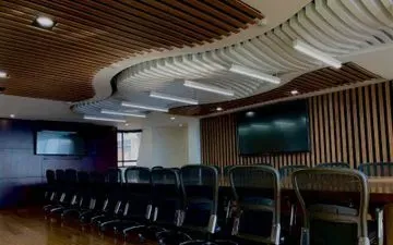 Sala de conferencias con paneles de madera, techo curvo con luces y sillas negras alrededor de una mesa larga.