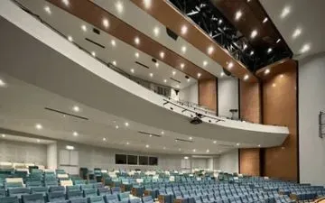 Interior del auditorio con filas de asientos azules, un balcón curvo e iluminación cenital. Las paredes son de colores neutros, con paneles marrones acentuados.