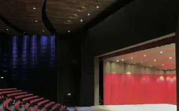 Un escenario de teatro vacío con cortinas rojas. Asientos rojos frente al escenario, con iluminación tenue y un techo oscuro y curvo.