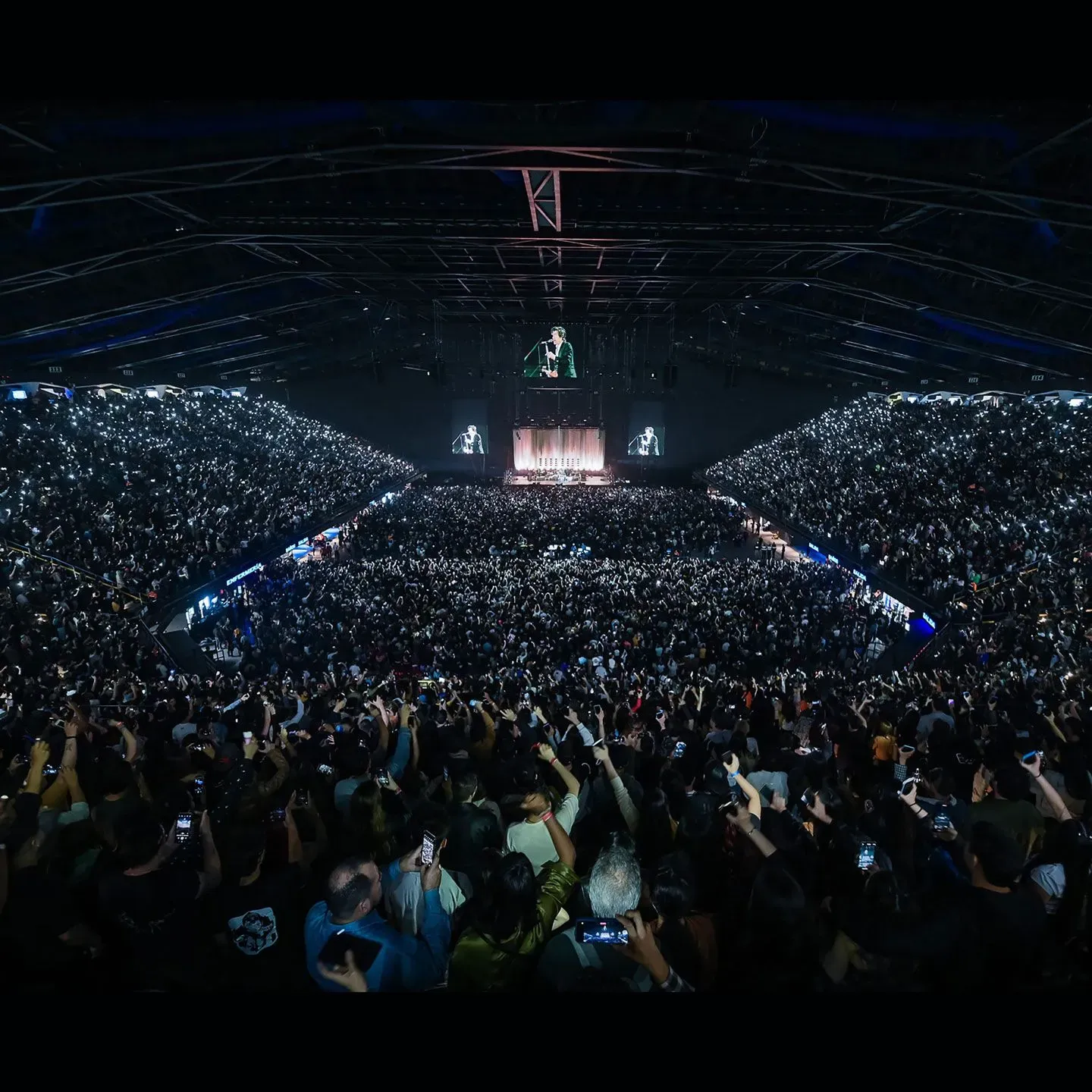Una gran sala de conciertos llena de gente sosteniendo sus teléfonos y con luces brillando hacia un escenario con un artista.