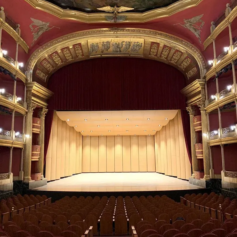 Interior de un gran teatro con detalles en rojo y dorado, un gran escenario y filas de asientos vacíos frente a él.