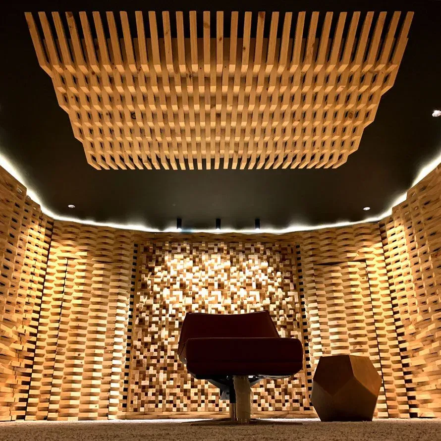 Estudio de grabación acústica con paredes y techo de listones de madera, una silla roja y un taburete geométrico.