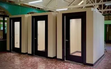 Tres cabinas insonorizadas con puertas negras y paneles de cristal, en una sala con suelo de baldosas rojas y blancas.