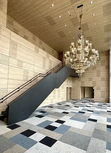 Amplio interior con una gran escalera, lámpara de araña y alfombra con motivos geométricos. Paredes y techo beige.