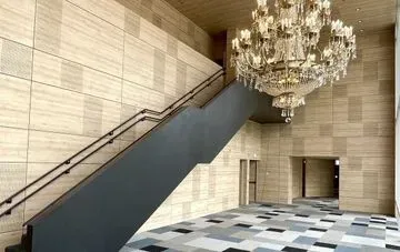 Fotografía interior de la entrada de un edificio con una escalera gris oscuro, una lámpara de araña y suelo de baldosas estampadas. Las paredes están cubiertas de bloques rectangulares de color claro.