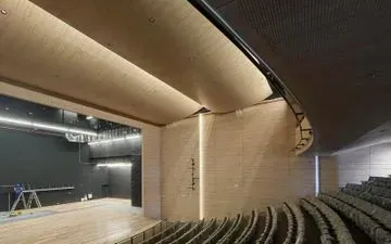 Interior de un teatro en construcción. Los paneles del techo de color madera clara se curvan sobre el escenario y la zona de asientos, con filas de asientos visibles a la derecha.