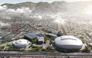 Vista aérea de un complejo deportivo moderno con grandes cúpulas blancas y otros edificios, frente a un paisaje urbano en expansión y montañas.