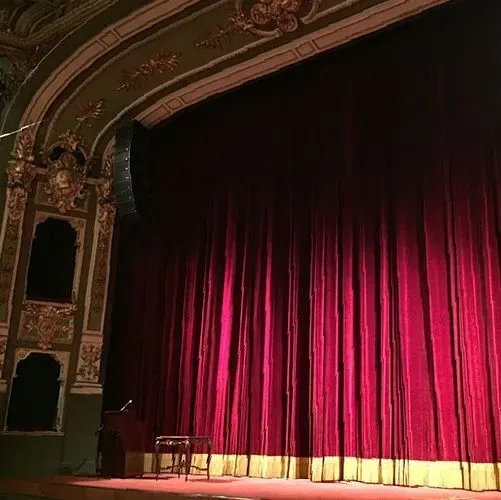 Escenario con cortinas de terciopelo rojo cerradas, detalles arquitectónicos ornamentados en oro y verde. Taburete de madera vacío y atril de micrófono sobre el escenario.