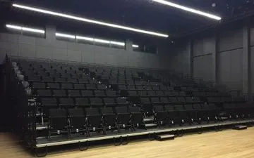 Filas de butacas negras en un auditorio moderno de paredes oscuras. Suelo de madera en primer plano, luces brillantes en el techo.