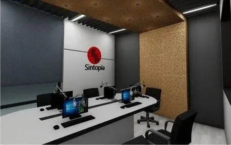 Interior de un estudio de radio con una mesa blanca con computadoras y micrófonos. La pared tiene un logotipo y la sala cuenta con paneles acústicos.