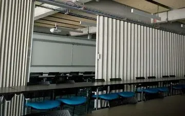 Un tabique blanco, largo y plegable separa dos habitaciones en una oficina moderna. El lado derecho cuenta con mesas y sillas azules.