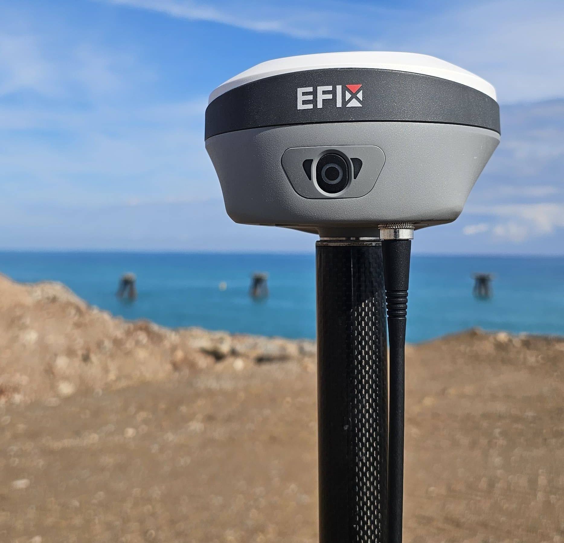 Un dispositivo GPS EFIx gris y blanco montado en un poste, frente a un océano y un cielo azules.