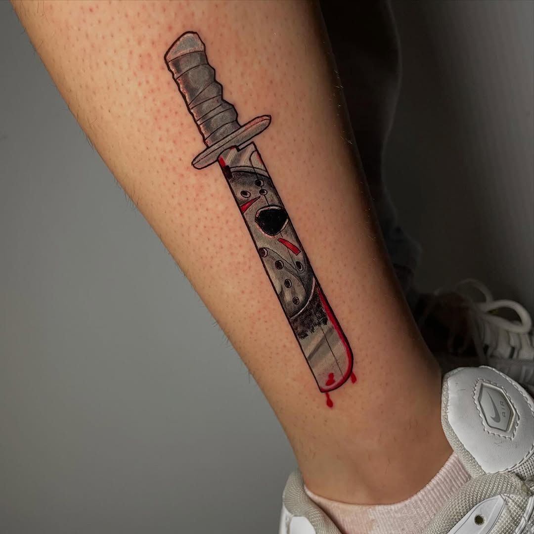 Un tatuaje en la pierna que representa una daga con mango gris y detalles rojos, cerca de una zapatilla blanca.