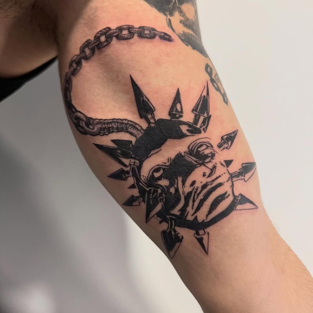 Tatuaje negro de un sol con cara de enfado, cadena y rayos en la parte superior del brazo.