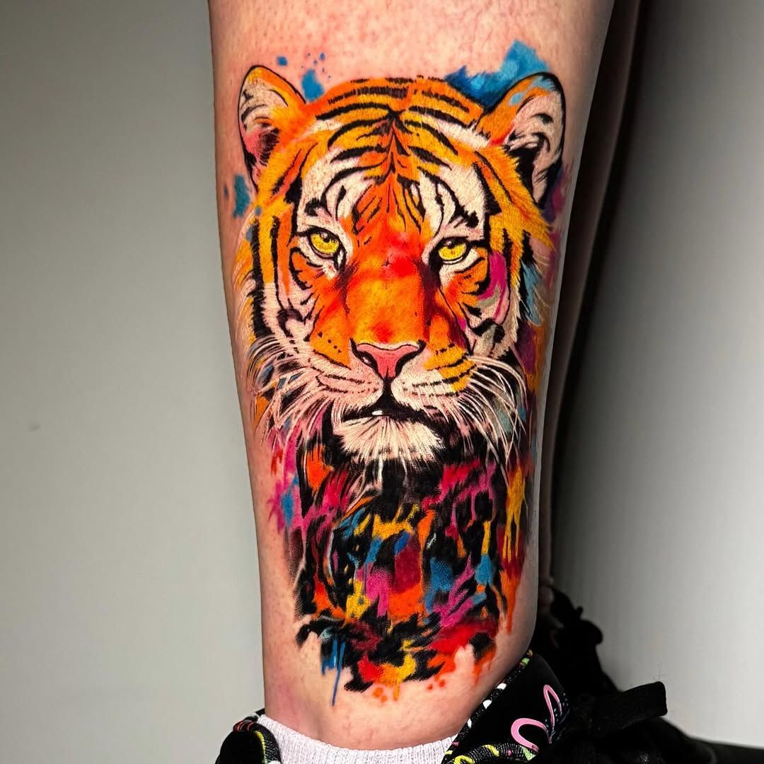 Tatuaje de tigre colorido en un antebrazo, con salpicaduras de color naranja brillante, negro y rosa a su alrededor.
