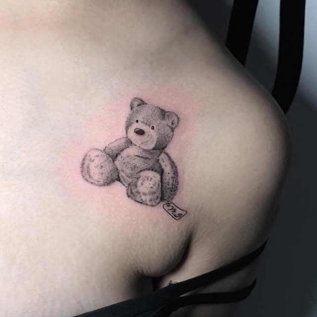 Pequeño tatuaje de un osito de peluche en un hombro con sombreado suave y piel rosada a su alrededor.