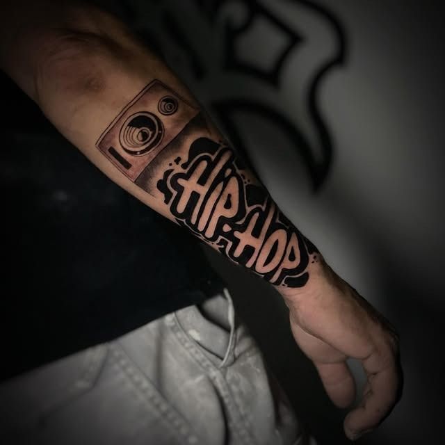 Primer plano de un antebrazo con un tatuaje de hip hop negro y cobre, junto a una camisa oscura y unos vaqueros claros.