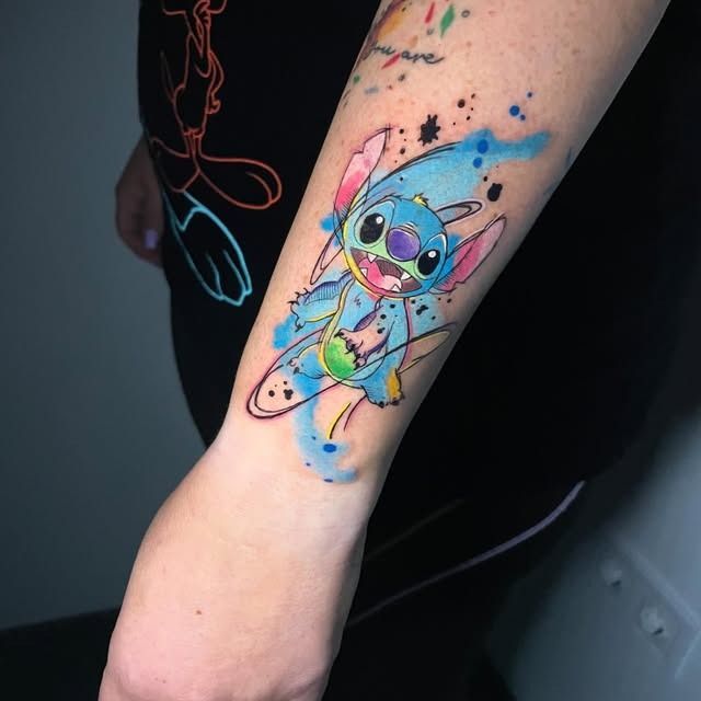Tatuaje colorido de Stitch con salpicaduras de acuarela en el antebrazo de una persona.