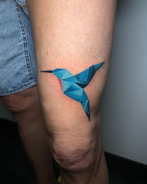 Tatuaje geométrico de un pájaro azul en la parte superior de la pierna, cerca de la rodilla.