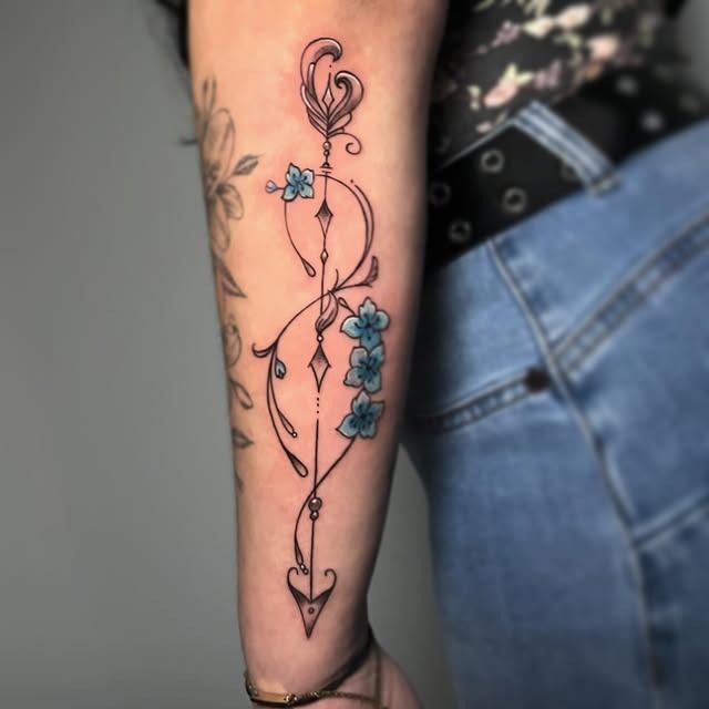 Primer plano de un antebrazo con un delicado tatuaje de una enredadera negra y pequeñas flores azules.