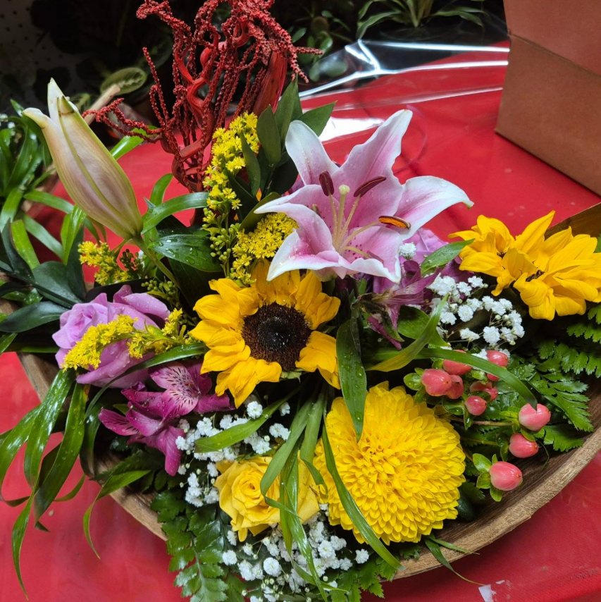 Composition florale colorée dans un bol en bois ; comprend des tournesols, des lys et d'autres fleurs assorties.