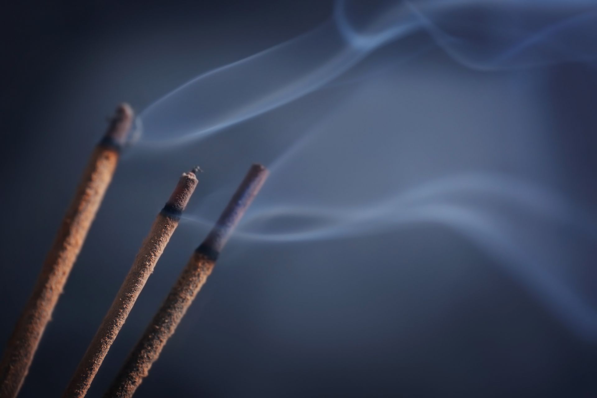 Trois bâtons d'encens brûlants avec des volutes de fumée bleue sur un fond sombre.