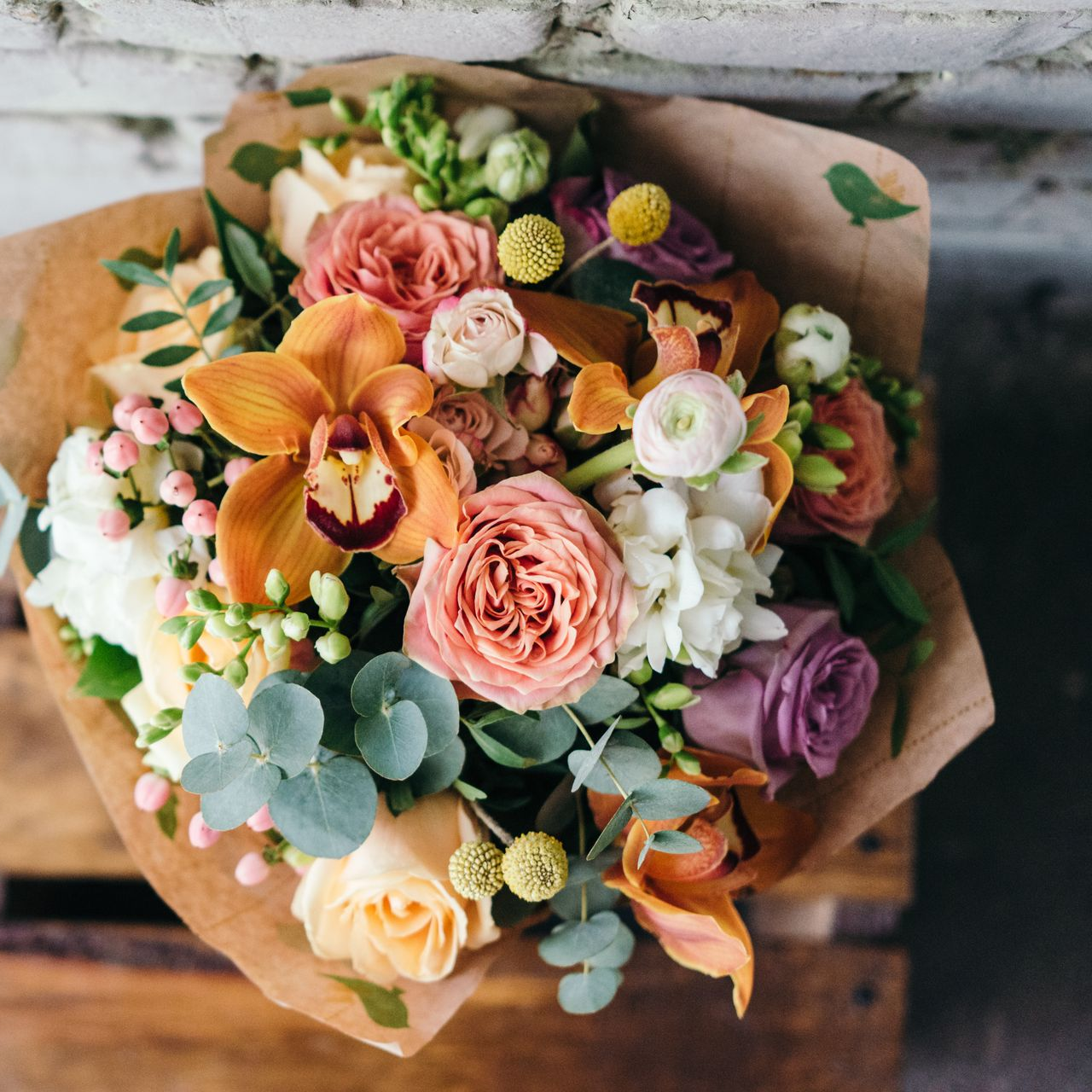 Bouquet de fleurs orange, roses et violettes enveloppées dans du papier brun, posées sur du bois.