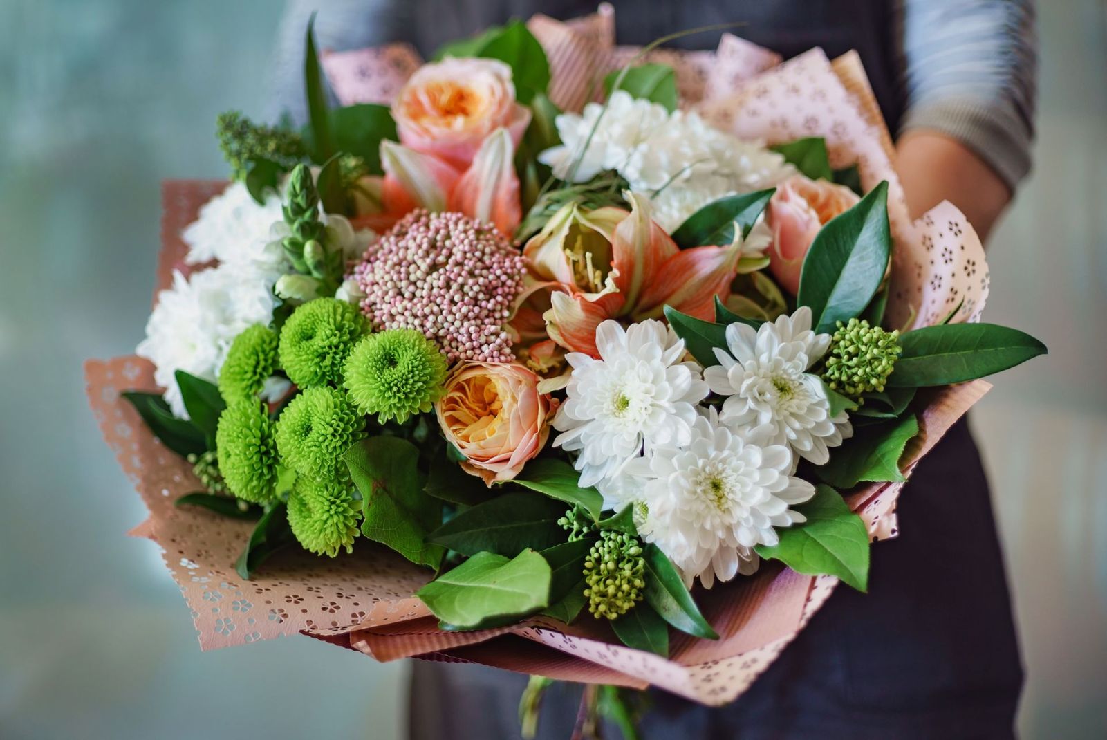 Femme tenant un bouquet de fleurs colorées, notamment des roses, des chrysanthèmes et de la verdure, enveloppés dans du papier décoratif.