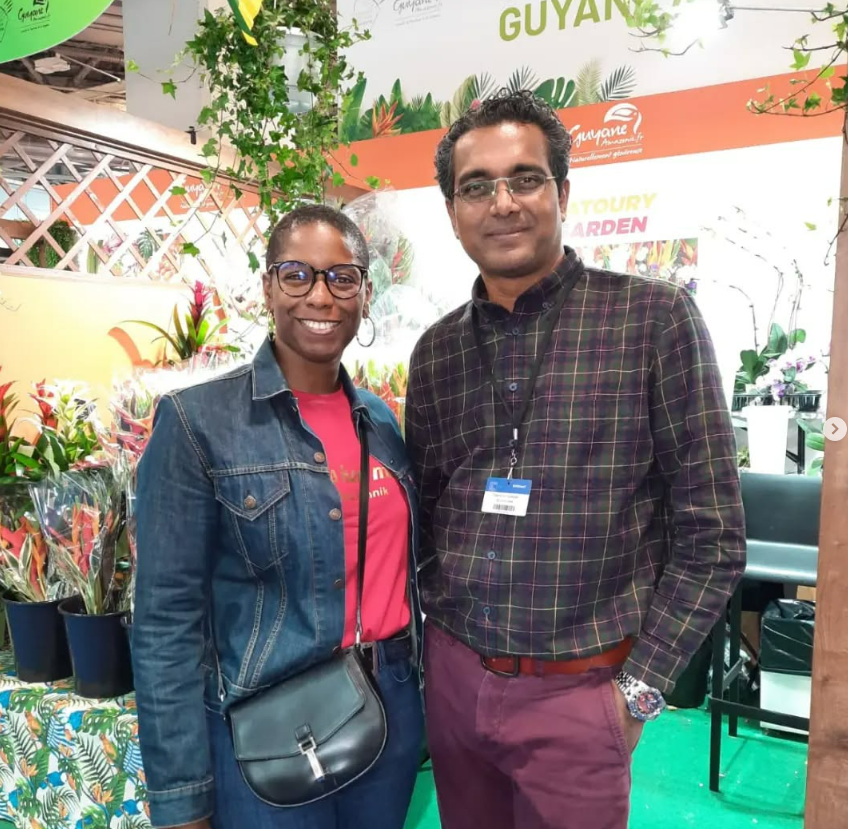 Deux personnes souriant devant une exposition de plantes. Une femme en veste en jean, un homme en chemise à carreaux.