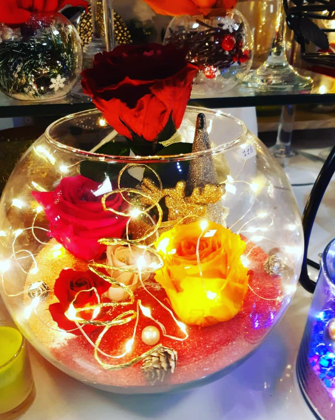 Un centre de table en verre avec des roses colorées, des guirlandes lumineuses et des éléments décoratifs.