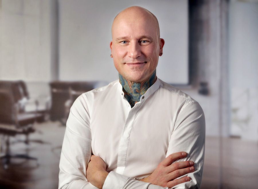 Ein kahlköpfiger Mann mit einer Tätowierung am Hals lächelt mit verschränkten Armen.